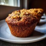 Close-up of a freshly baked, golden brown banana nut muffin topped with walnuts, sitting on a white plate.