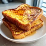 Two thick slices of golden brown easy french toast stacked on a white plate, drizzled with syrup.