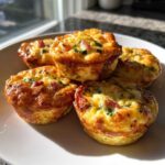 A close-up of five golden brown Egg Muffin Cups piled on a white plate, baked with visible ham and herbs.