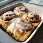 Four gooey, freshly baked cinnamon rolls topped with thick white icing in a white baking dish.