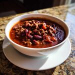 A close-up of a white bowl filled with rich, dark red crockpot chili featuring ground meat and kidney beans.