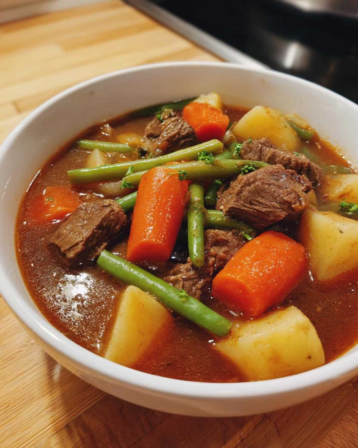 Close-up of a white bowl filled with rich vegetable beef soup, featuring large chunks of beef, carrots, potatoes, and green beans.