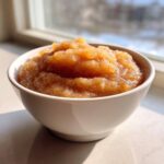 A close-up of chunky, homemade applesauce served in a white bowl, backlit by a window.
