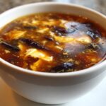 A close-up view of a white bowl filled with steaming hot and sour soup recipe, showing tofu and dark mushrooms.