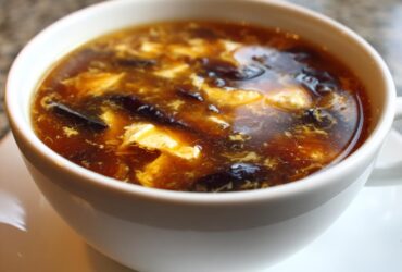 A close-up view of a white bowl filled with steaming hot and sour soup recipe, showing tofu and dark mushrooms.