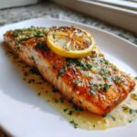 Close-up of a perfectly cooked baked salmon recipe fillet topped with garlic, herbs, and a charred lemon slice.