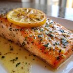 Close-up of a perfectly cooked baked salmon recipe fillet topped with garlic, herbs, and a lemon slice.