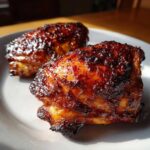 Close-up of two pieces of juicy bbq chicken with a dark, caramelized glaze resting on a white plate.