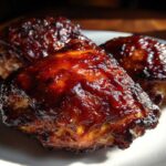 Three pieces of incredibly juicy bbq chicken coated in a thick, dark, glossy sauce, resting on a white plate.