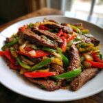 Close-up of perfectly cooked steak fajitas featuring sliced steak, red and green bell peppers, and onions on a white plate.