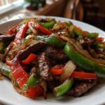 Close-up of juicy steak fajitas mixture with sautéed green peppers, red peppers, and onions on a white plate.