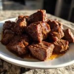A pile of tender, glazed, bite-sized pieces of crockpot steak served on a white plate.