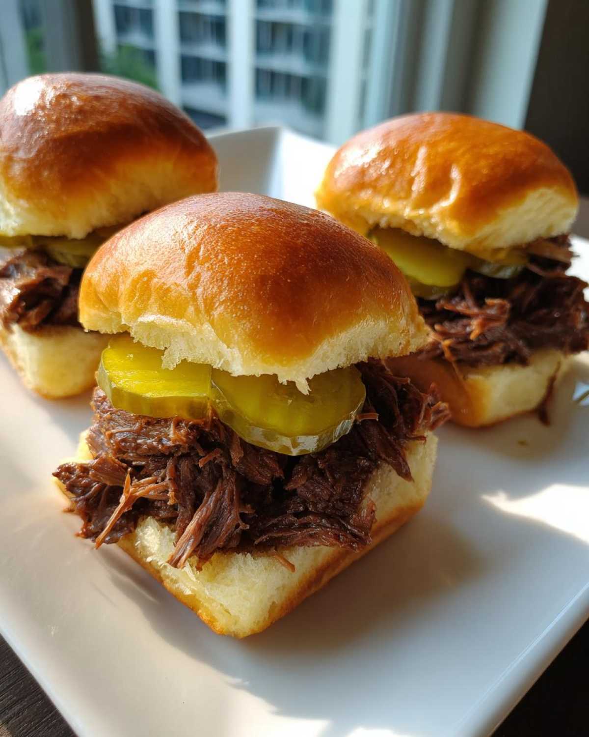 Three delicious mississippi pot roast sliders piled high with shredded beef and pickle slices on brioche buns.