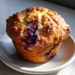 Close-up of a perfectly baked, golden-brown blueberry muffin with visible burst blueberries and a sugary crust.