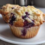 Close-up of a freshly baked, moist blueberry muffin topped with a golden crumb topping, sitting on a white plate.