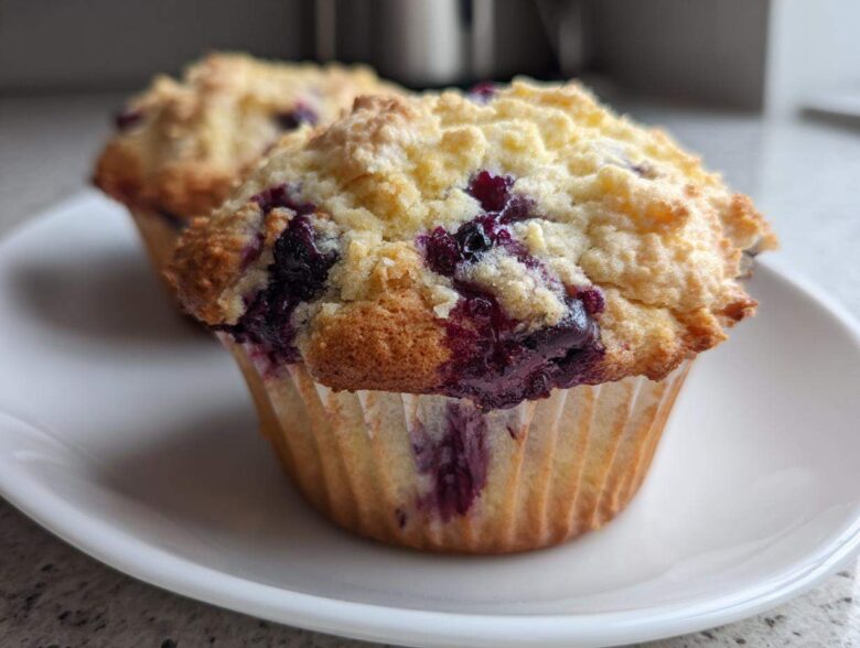 Close-up of a freshly baked, moist blueberry muffin topped with a golden crumb topping, sitting on a white plate.