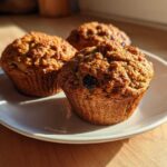 Three golden brown, moist bran muffins with visible raisins, sitting on a white plate in warm sunlight.