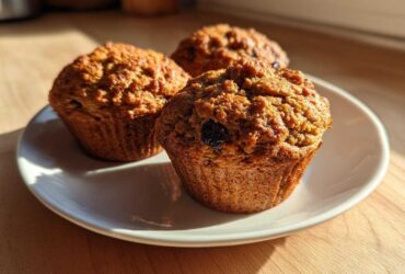 Three golden brown, moist bran muffins with visible raisins, sitting on a white plate in warm sunlight.