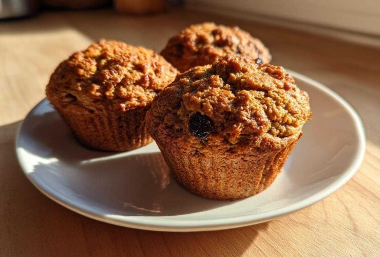 Three golden brown, moist bran muffins with visible raisins, sitting on a white plate in warm sunlight.