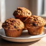 Three golden brown, moist bran muffins with visible raisins, sitting on a light speckled plate.