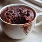 A close-up of a steaming, moist chocolate mug cake topped with chocolate chips inside a white mug.
