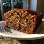 A close-up of a thick slice of moist date nut bread, rich with visible nuts and raisins, sitting on a white plate.