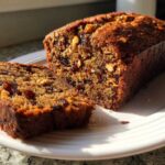 A close-up of a freshly baked loaf of date nut bread, with one thick slice cut and resting beside it on a white plate.