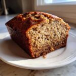 A close-up of a moist slice of sugar free banana bread with a dark, caramelized crust, resting on a white plate.