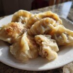 A plate of golden-brown, pan-fried Traditional Chinese Dumplings, one cut open showing the filling.