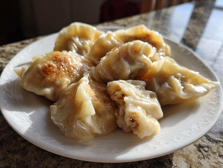 A plate of golden-brown, pan-fried Traditional Chinese Dumplings, one cut open showing the filling.