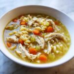 Close-up of a white bowl filled with hot penicillin soup featuring shredded chicken, carrots, celery, and rotini pasta.