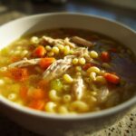 Close-up of a warm bowl of penicillin soup featuring shredded chicken, elbow macaroni, and bright orange carrots.