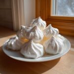 A pile of freshly piped, bright white meringue cookies stacked on a small white plate near a sunny window.