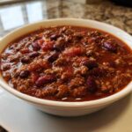 A close-up of a white bowl filled with rich, hearty crockpot chili featuring ground meat and kidney beans.