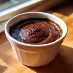 A close-up of rich, dark homemade chocolate pudding swirled in a small white ramekin, sitting on a wooden surface.