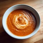 Overhead view of a bowl of rich, orange-red tomato bisque, garnished with a swirl of cream.