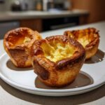 Three golden brown, perfectly risen Yorkshire puddings sitting on a white plate, showing their deep, hollow centers.