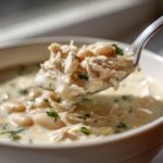 A spoonful of creamy crockpot white chicken chili featuring shredded chicken and white beans being lifted from a bowl.