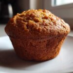 A single, golden-brown honey bran muffin sitting on a white plate, highlighted by natural light.