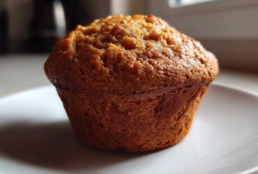 A single, golden-brown honey bran muffin sitting on a white plate, highlighted by natural light.
