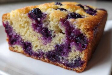 Close-up of a moist slice of blueberry banana bread showing bright purple blueberries baked into the yellow crumb.