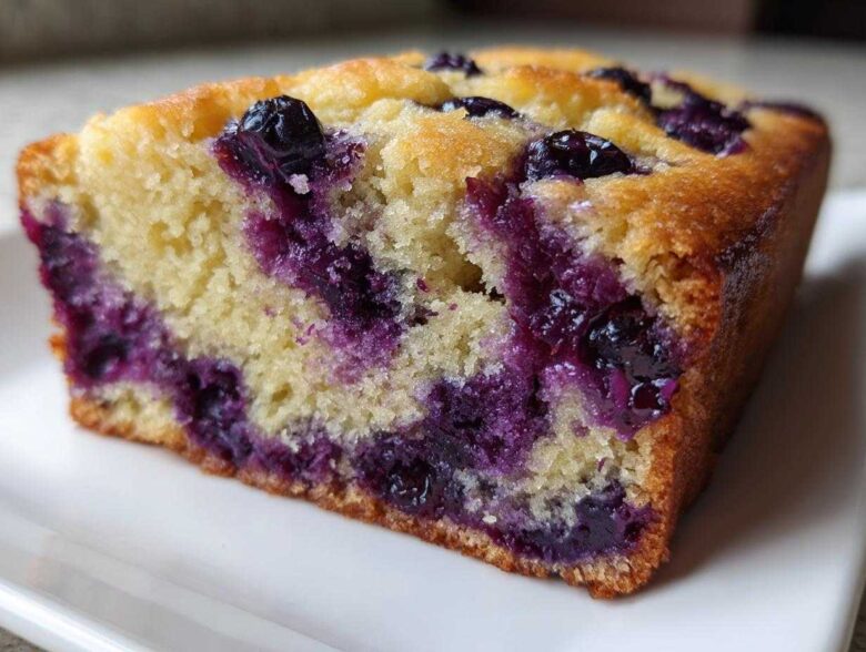 Close-up of a moist slice of blueberry banana bread showing bright purple blueberries baked into the yellow crumb.