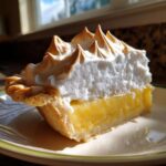 A close-up slice of bright yellow lemon meringue pie showing the tall, toasted meringue peaks.