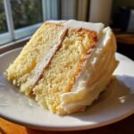 Close-up of a moist slice of vanilla cake with thick white frosting, bathed in sunlight.