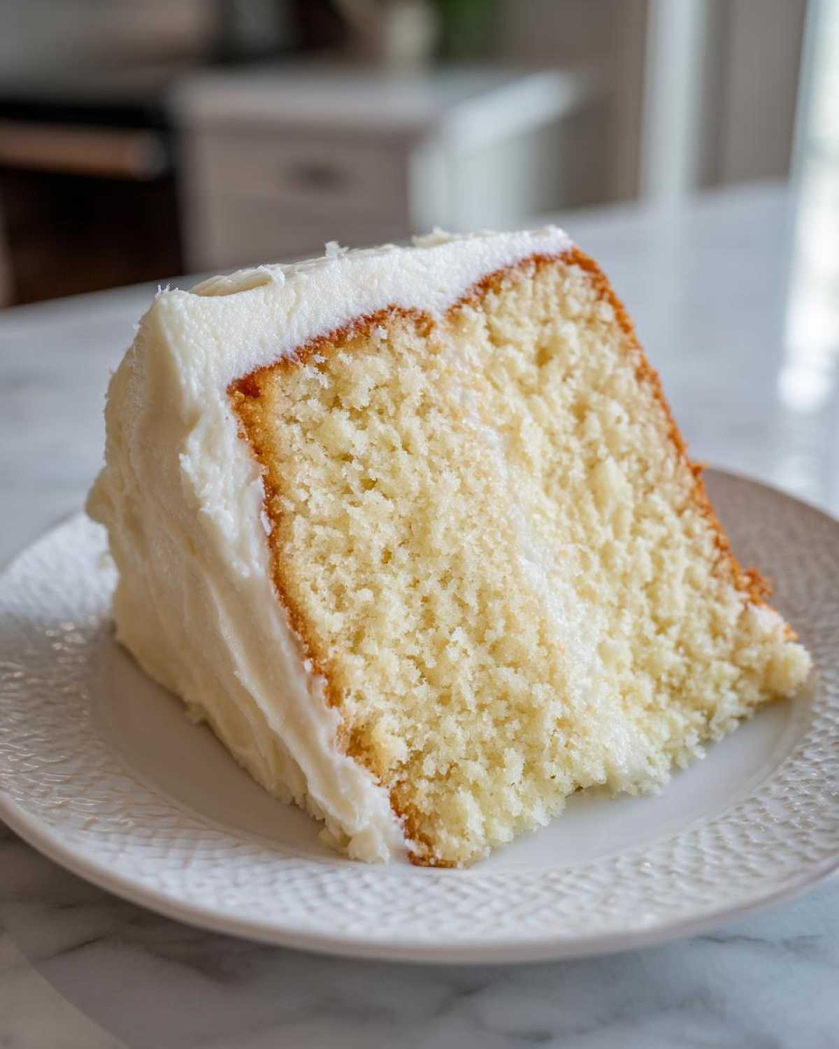 Close-up of a moist slice of vanilla cake recipe, topped with thick white buttercream frosting, served on a white plate.