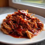 A close-up of tender, saucy slow cooker pulled pork piled high on a white plate.