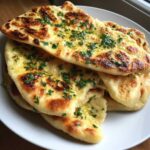 A stack of freshly made, soft naan bread brushed with melted butter and topped with chopped green herbs.