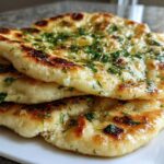 Close-up of a stack of three soft, golden-brown naan bread pieces brushed with butter and sprinkled with fresh parsley.
