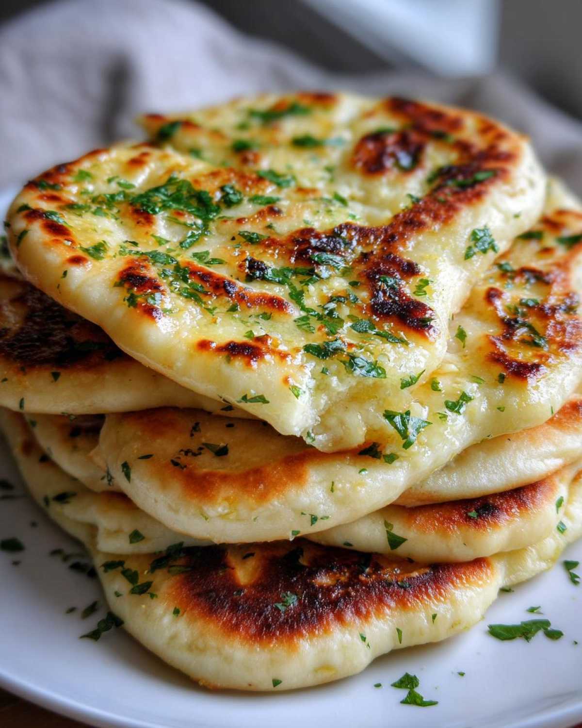 A close-up stack of freshly made, soft naan bread brushed with butter and topped with chopped green herbs.