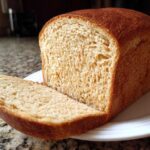 A freshly baked loaf of soft whole wheat bread with one thick slice cut and resting against the main loaf on a white plate.